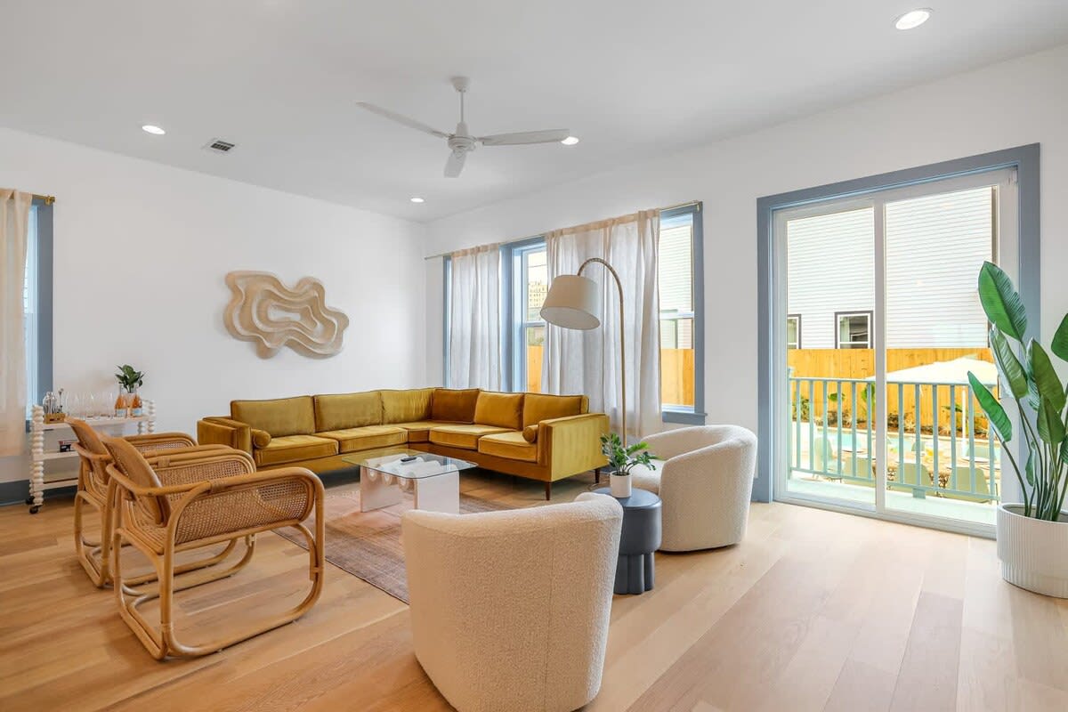Sunlit modern living room with a mustard yellow L-shaped sofa, rattan accent chairs, two cream boucle lounge chairs, glass coffee table, light oak floors, tall arc floor lamp, large windows and sliding glass door leading to a small balcony, and potted greenery.