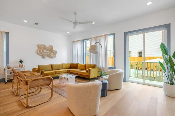 Sunlit modern living room with a mustard yellow L-shaped sofa, rattan accent chairs, two cream boucle lounge chairs, glass coffee table, light oak floors, tall arc floor lamp, large windows and sliding glass door leading to a small balcony, and potted greenery.