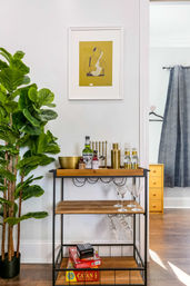 Bright modern interior corner with a wooden-and-metal home bar cart stocked with liquor bottles, a brass cocktail shaker, martini glasses and hanging stemware, a tall fiddle leaf fig plant at left, a yellow saxophone print framed above, and board games (including Catan) on the bottom shelf — cozy game-night ready setup.