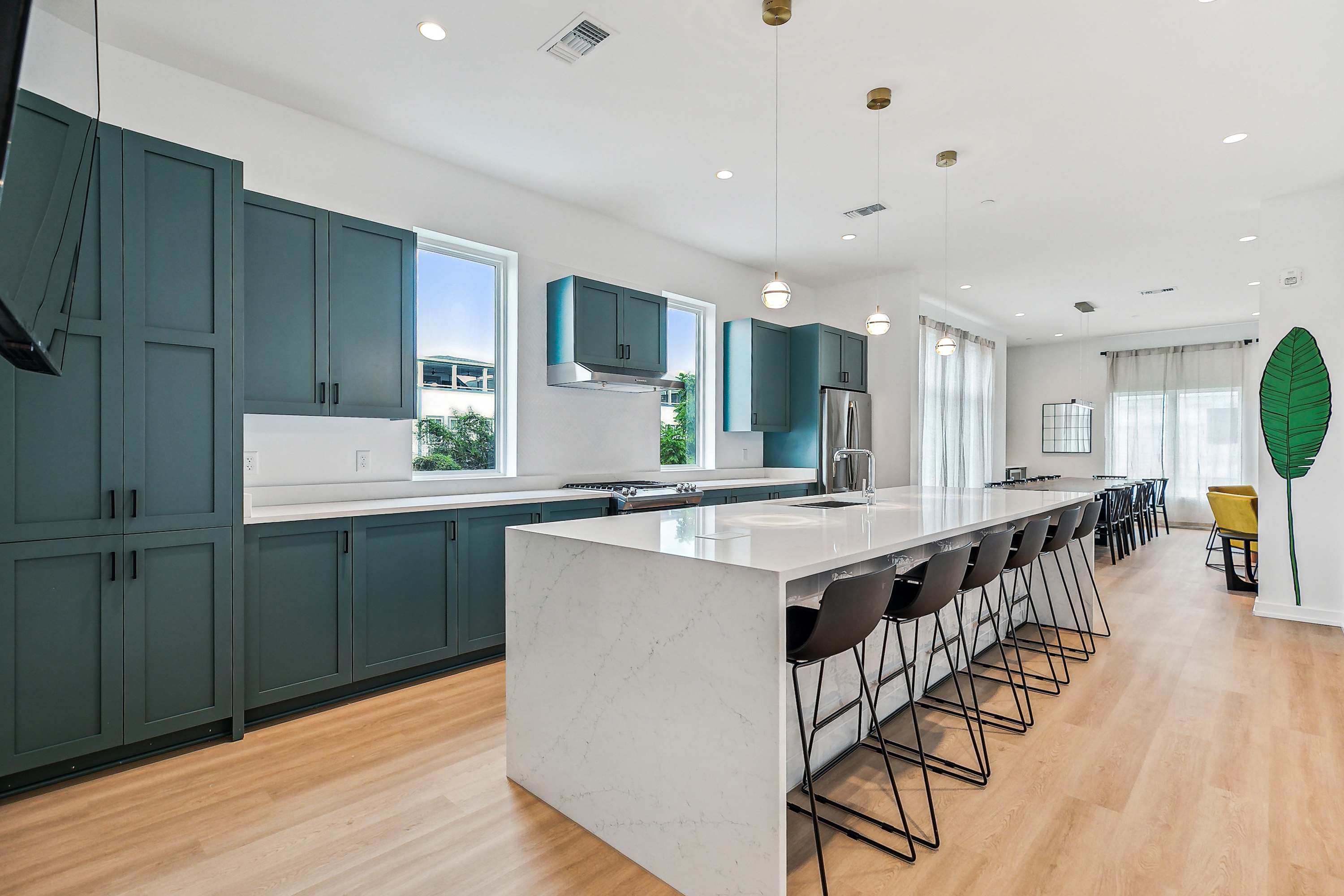 Modern open-concept kitchen with long white waterfall island, teal shaker cabinets, black bar stools, pendant lights, light wood floors and adjacent dining area.