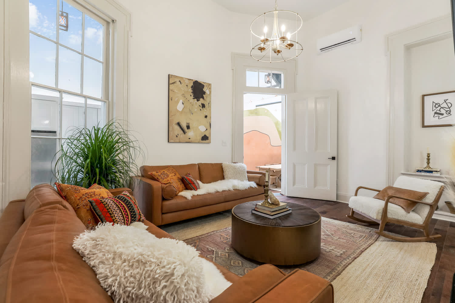 Sunlit boho-chic living room with caramel leather sofas, round wooden coffee table, shag throws, patterned pillows, large window, indoor plant and modern chandelier