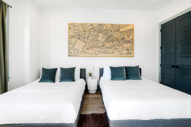 Bright modern guest bedroom with two side-by-side beds dressed in white linens and teal velvet pillows, round white bedside table with a small plant and lamp, large playful line-art framed print above the beds, dark paneled closet doors and light wood floor.