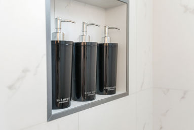 Modern marble shower niche holding three sleek black pump dispensers labeled shampoo, body soap, and conditioner.