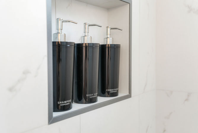 Modern marble shower niche holding three sleek black pump dispensers labeled shampoo, body soap, and conditioner.