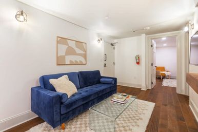 Bright modern apartment living room with a blue velvet sofa, glass coffee table, shag rug, hardwood floors, minimalist wall art and cozy lighting.