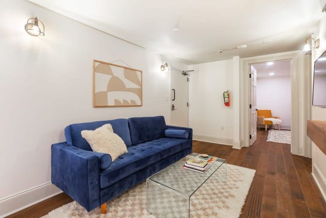 Bright modern apartment living room with a blue velvet sofa, glass coffee table, shag rug, hardwood floors, minimalist wall art and cozy lighting.