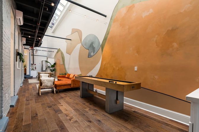 Industrial loft-style lounge with skylight, large abstract wall mural, dark hardwood floors, a wooden shuffleboard table, burnt-orange sectional sofa and cozy sheepskin chairs.