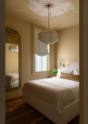 Cozy neutral-toned bedroom in a warm city apartment with a plush bed, linen throw, geometric pendant light, tall decorative mirror, roman shade window, and bedside plant.