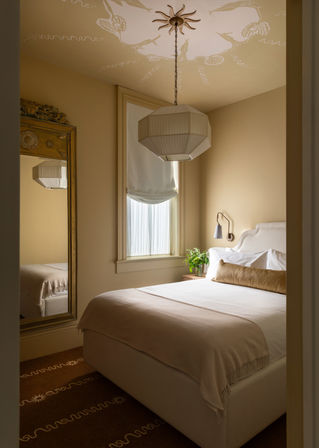 Cozy neutral-toned bedroom in a warm city apartment with a plush bed, linen throw, geometric pendant light, tall decorative mirror, roman shade window, and bedside plant.
