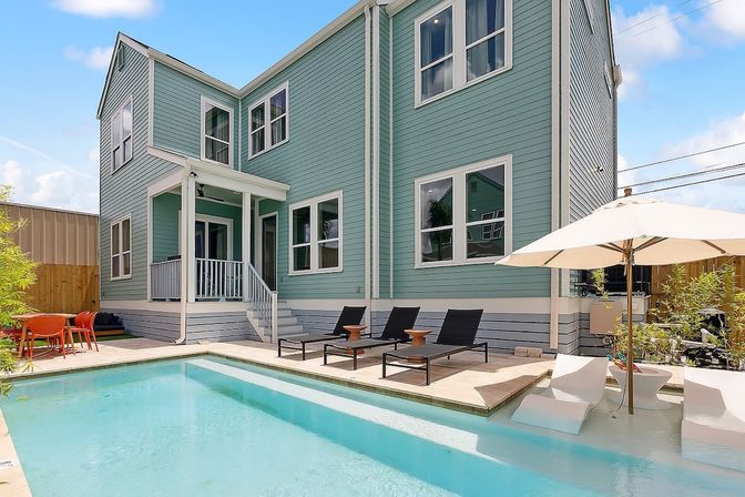 Sunlit backyard pool beside a two-story teal house with patio dining, black loungers, and a white umbrella