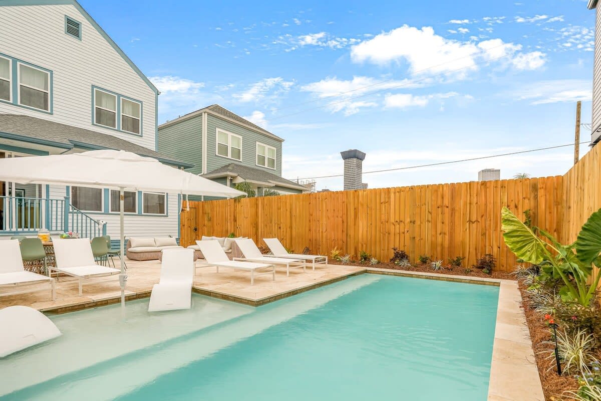 Backyard swimming pool with shallow tanning ledge, white lounge chairs and umbrella on a stone patio beside two-story homes and wooden privacy fence