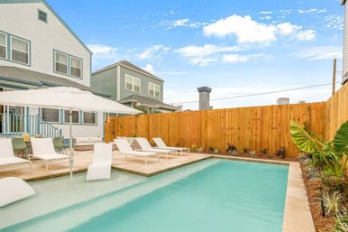 Backyard swimming pool with shallow tanning ledge, white lounge chairs and umbrella on a stone patio beside two-story homes and wooden privacy fence