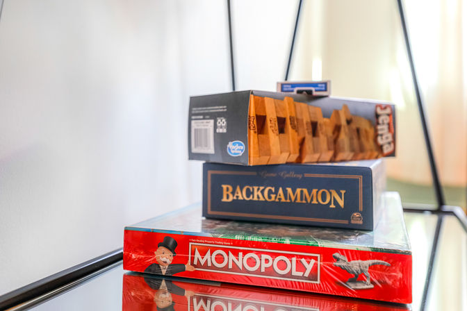 Stack of classic board games — Monopoly, Backgammon and a wooden block stacking game — neatly piled on a glass side table in a bright room
