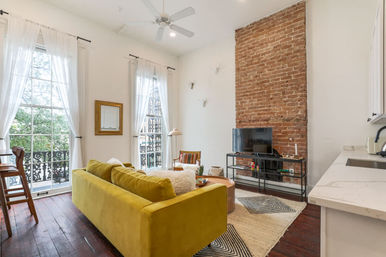 Bright city apartment living room with mustard velvet sofa, exposed brick chimney, tall windows with white curtains and wrought-iron balcony railings, TV on metal stand, hardwood floors and open kitchen counter.