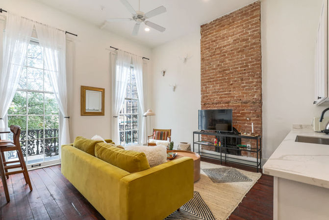 Bright city apartment living room with mustard velvet sofa, exposed brick chimney, tall windows with white curtains and wrought-iron balcony railings, TV on metal stand, hardwood floors and open kitchen counter.