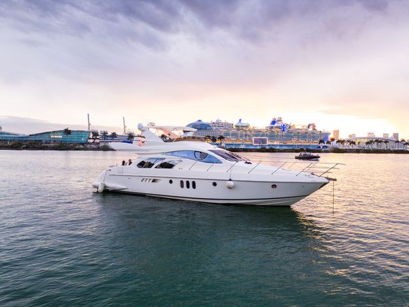 Luxury white motor yacht in a calm tropical harbor with a large cruise ship, palm-lined waterfront and pastel sunset sky.