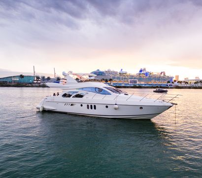 Luxury white motor yacht in a calm tropical harbor with a large cruise ship, palm-lined waterfront and pastel sunset sky.