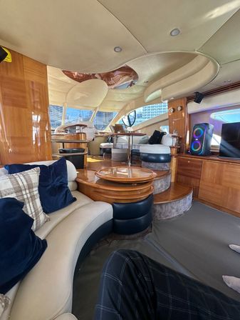 Sunlit luxury yacht salon with curved cream leather seating, navy pillows, glossy wood tables and stepped teak flooring leading to the helm, large windows framing a marina and city skyline, LED speaker and sculpted ceiling details.