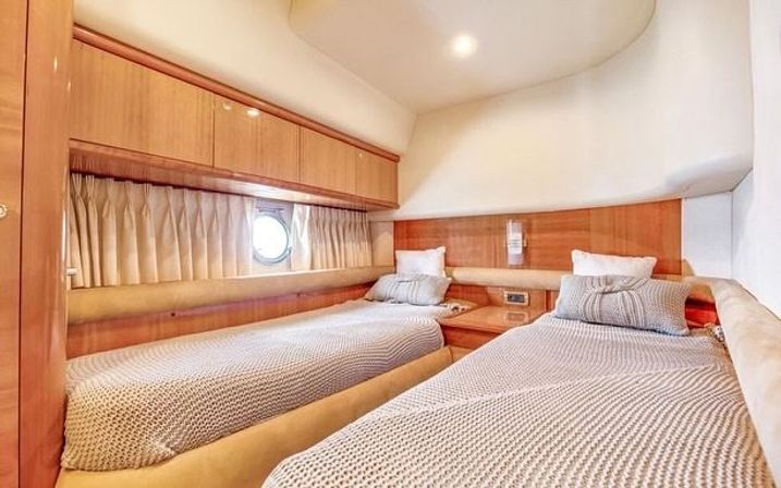 Cozy yacht cabin with twin beds, beige knit throws, teak wood cabinetry, round porthole and soft overhead lighting — a bright nautical guest stateroom.