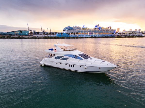 White luxury yacht cruising a calm harbor at sunset, bathed in golden light with a large cruise ship, palm‑lined waterfront and distant city skyline in the background.
