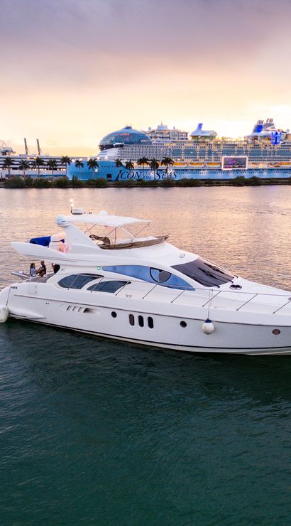 White luxury yacht cruising a calm harbor at sunset, bathed in golden light with a large cruise ship, palm‑lined waterfront and distant city skyline in the background.