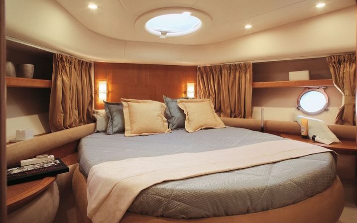 Cozy luxury yacht cabin interior with round porthole and overhead hatch, circular bed dressed in blue-gray linens, beige throw and pillows, warm wood paneling and soft ambient lighting.