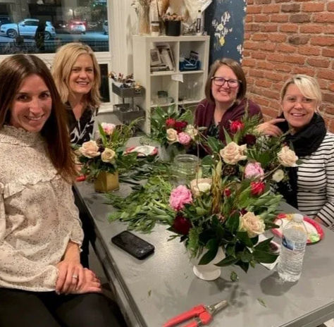 Four smiling women at a cozy indoor flower-arranging workshop, seated around a table with rose bouquets, greenery, clippers, vases, a phone and water bottle against an exposed brick wall.