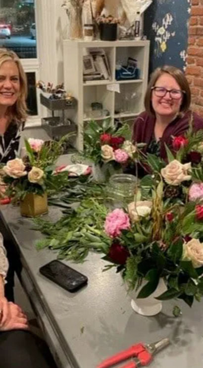 Four smiling women at a cozy indoor flower-arranging workshop, seated around a table with rose bouquets, greenery, clippers, vases, a phone and water bottle against an exposed brick wall.