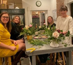 Four women smiling at a flower-arranging workshop in a cozy studio, creating rose and greenery arrangements on a worktable with pruning shears, a 'Fresh Cut Flowers' sign and wall clock in the background.