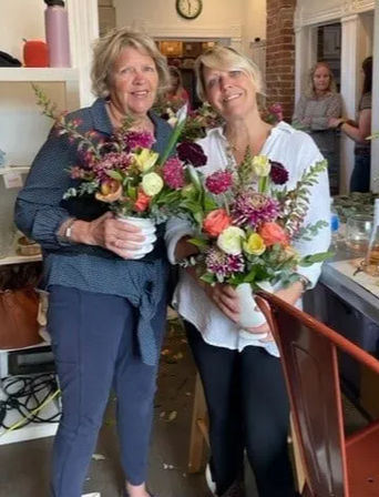 Two smiling women holding vibrant mixed bouquets inside a cozy local flower shop with rustic decor and seasonal floral arrangements.