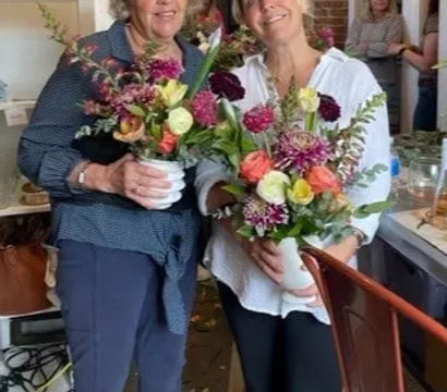 Two smiling women holding vibrant mixed bouquets inside a cozy local flower shop with rustic decor and seasonal floral arrangements.