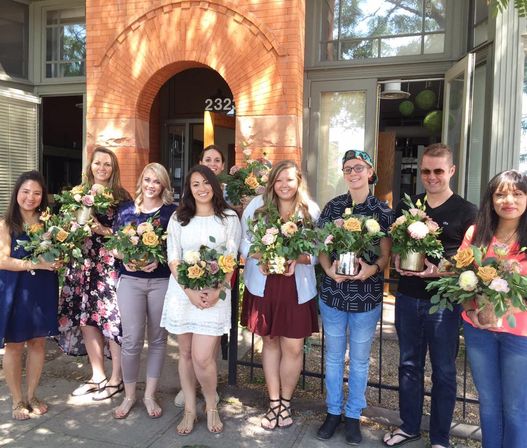 Group of people holding fresh floral bouquets outside a brick building with an arched entrance — outdoor flower-arranging class on a sunny day.