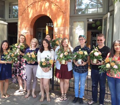 Group of people holding fresh floral bouquets outside a brick building with an arched entrance — outdoor flower-arranging class on a sunny day.