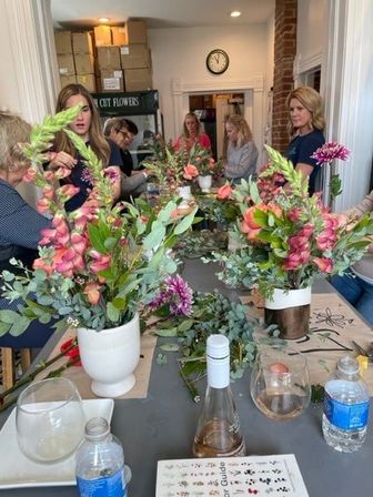 Hands-on floral arranging workshop in a bright studio with participants creating pink snapdragon, coral rose and eucalyptus bouquets in white vases on a communal table.