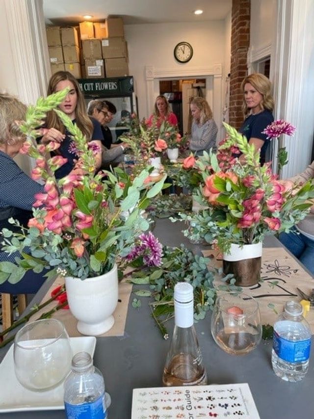Hands-on floral arranging workshop in a bright studio with participants creating pink snapdragon, coral rose and eucalyptus bouquets in white vases on a communal table.