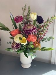 Cheerful mixed floral bouquet in a white ceramic vase on a tabletop — coral roses, yellow tulip, cream ranunculus, purple chrysanthemums, eucalyptus and assorted greenery, indoor home-decor arrangement.