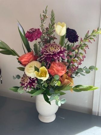 Cheerful mixed floral bouquet in a white ceramic vase on a tabletop — coral roses, yellow tulip, cream ranunculus, purple chrysanthemums, eucalyptus and assorted greenery, indoor home-decor arrangement.