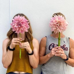 Two people side-by-side holding oversized pink dahlia blooms in front of their faces against a white wall, wearing casual tank tops, watches and rings — playful close-up portrait.