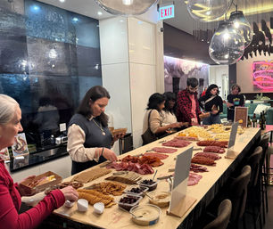 Large charcuterie grazing table in a modern indoor lounge with pendant lights — rows of cured meats, cheeses, crackers, olives and dips as guests serve themselves at a casual event