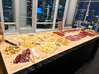 Long charcuterie buffet with assorted cubed cheeses, cheese logs, sliced salami and cured meats arranged on kraft paper with handwritten labels on a marble counter in a modern high‑rise lounge overlooking a nighttime city skyline.