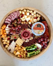 Party-ready round wooden charcuterie board with assorted cheeses (cubed, sliced, crumbled), folded cured meats, red grapes, sliced cucumber, nuts and cranberries, plus a small French onion dip container on a marble countertop.