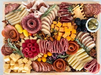 Gourmet charcuterie board on a wooden tray with assorted sliced and cubed cheeses, folded salami and prosciutto, pepperoni roses, blackberries and raspberries, green olives, mixed nuts and rosemary sprigs — party-ready appetizer platter.