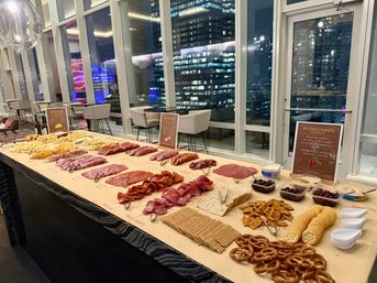 Long charcuterie spread of sliced cured meats, cheeses, crackers, pretzels and olives on a buffet table in a modern high-rise lounge with floor-to-ceiling windows and a nighttime city skyline view.
