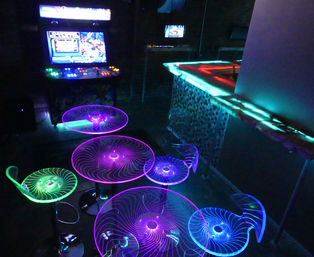 Neon-lit arcade bar interior with spiral LED tables and stools glowing purple, blue and green, a retro arcade machine and illuminated counter.