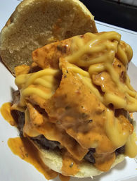 Close-up of a messy cheeseburger: beef patty topped with crunchy tortilla chips, drippy melted yellow cheese and spicy orange sauce on a soft bun.