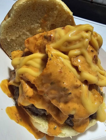 Close-up of a messy cheeseburger: beef patty topped with crunchy tortilla chips, drippy melted yellow cheese and spicy orange sauce on a soft bun.
