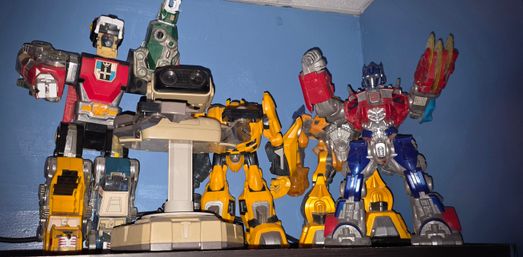 Shelf of colorful robot action-figure toys and collectibles—red, yellow, blue and beige mechs—posed on a wall shelf against a blue wall.