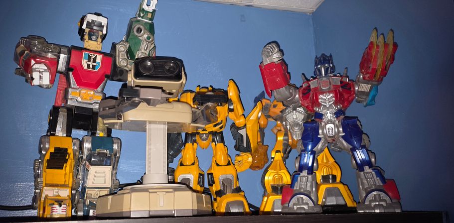 Shelf of colorful robot action-figure toys and collectibles—red, yellow, blue and beige mechs—posed on a wall shelf against a blue wall.