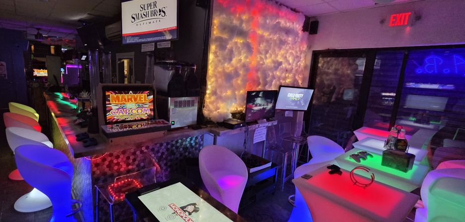 Neon-lit gaming lounge interior with colorful LED stools, glowing tables, arcade cabinet and multiple monitors displaying fighting and shooter games.