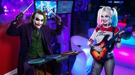 Cosplayers as Joker and Harley Quinn playing a Rock Band-style game in a neon-lit arcade, Joker on an electronic drum kit and Harley Quinn holding a toy guitar with colorful LED lights and game screens in the background.